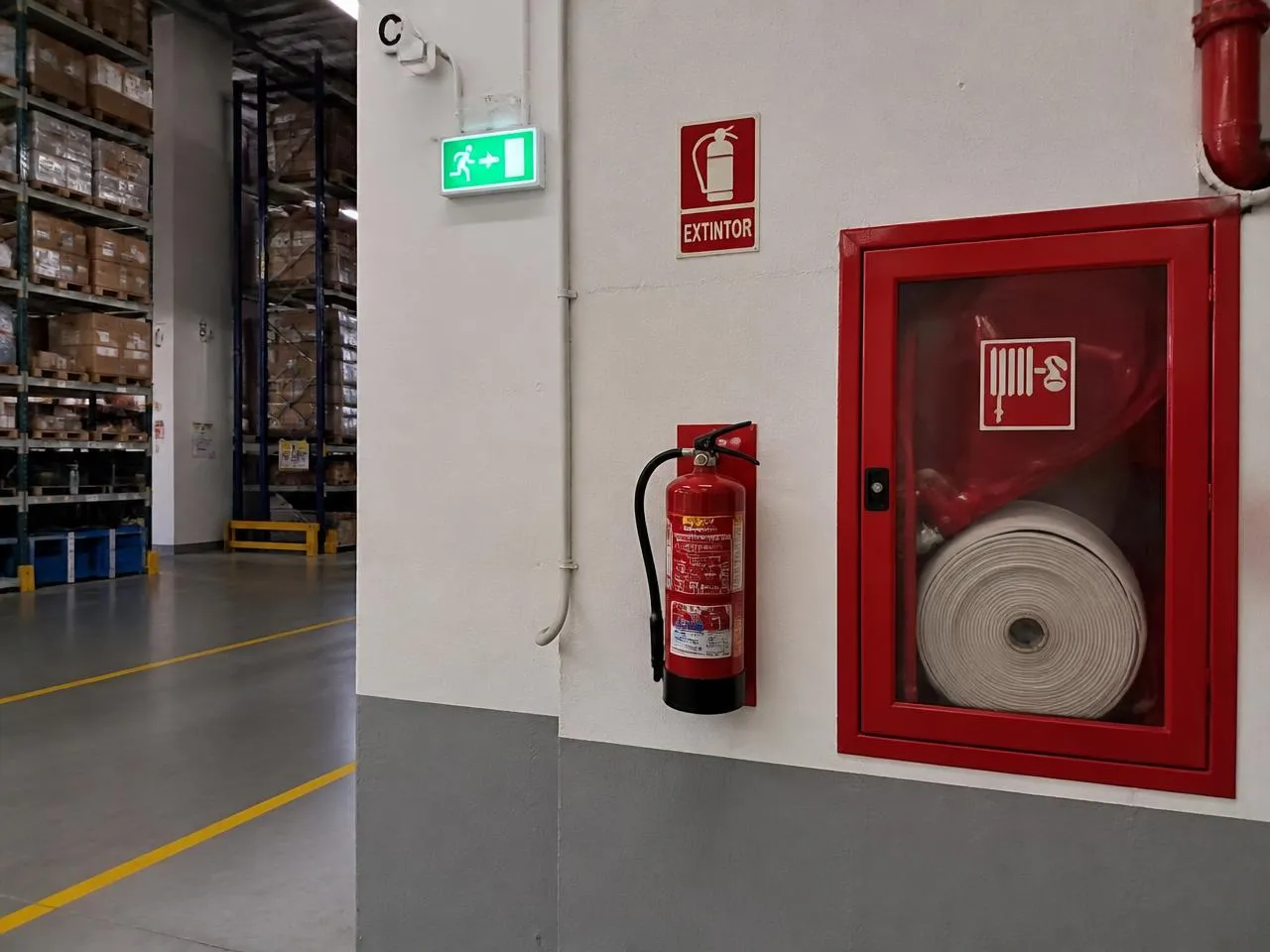 Parede de galpão industrial com abrigo de hidrante vermelho, extintor de incêndio, sinalização de extintor e placa de saída de emergência iluminada.
